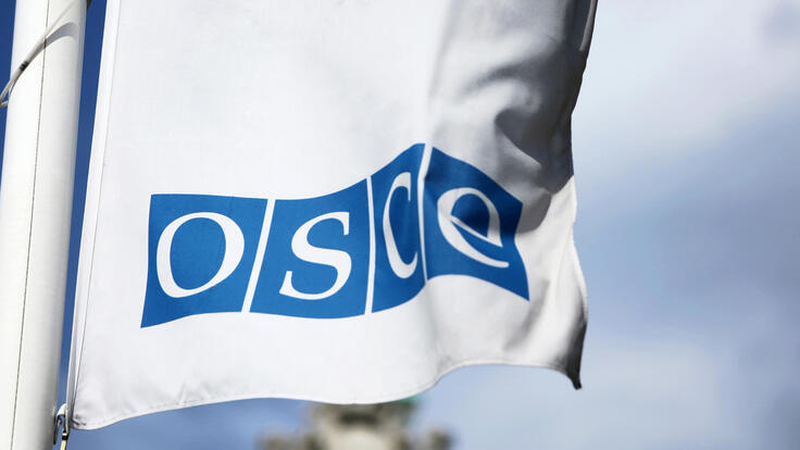 This is an image of a flag bearing the logo of the Organization for Security and Co-operation in Europe (OSCE). The OSCE logo features the acronym "OSCE" written in white letters within blue squares, arranged horizontally. The flag itself is white, and it is seen waving against a soft, cloudy blue sky in the background. The logo is a prominent element, signifying the organization's identity.