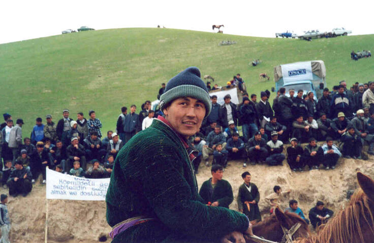 Man on horseback at an outdoor gathering with a crowd seated on a hillside and an OSCE truck in the background.