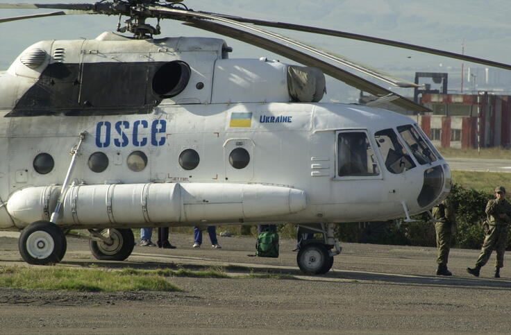 A white helicopter with the name of the OSCE and the Ukrainian flag standing on the tarmac of the Tbilisi military airport, with several soldiers standing nearby.