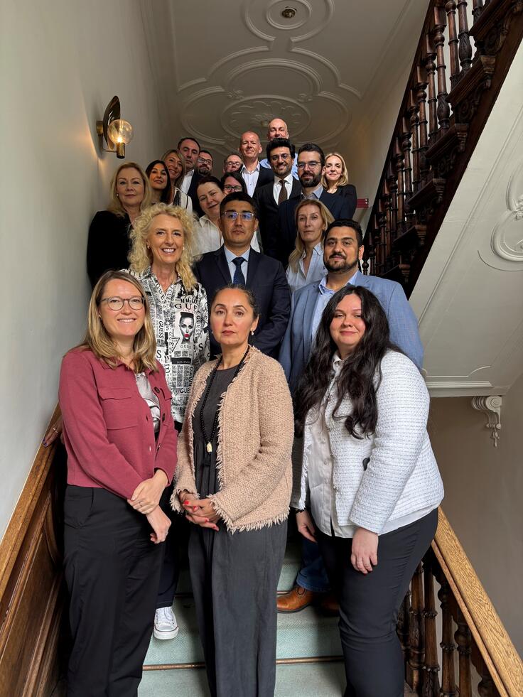 Group of people smiling, standing on a staircase with ornate railings in a well-lit interior.