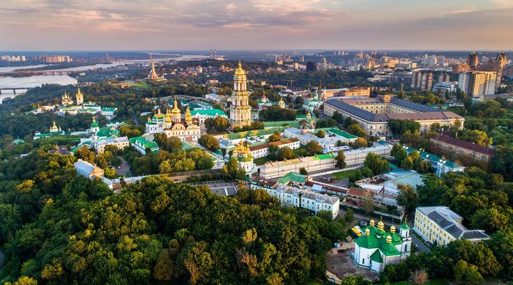 Aerial view of Kyiv Pechersk Lavra, golden domes, surrounded by green trees, cityscape in the background.