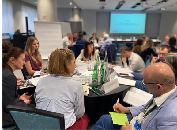 People seated around tables in a conference room, discussing and taking notes.