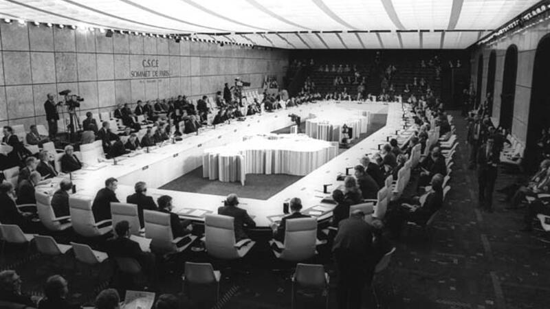 A black-and-white photograph of the CSCE Paris Summit, showing a large conference room with delegates seated around a U-shaped table arrangement. The backdrop includes the text 'CSCE Sommet de Paris,' and the space is filled with officials, media personnel, and observers.