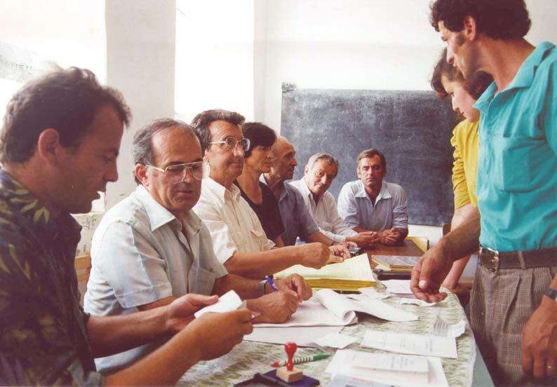 Election monitors seated around a table with papers, one standing; chalkboard in the background.
