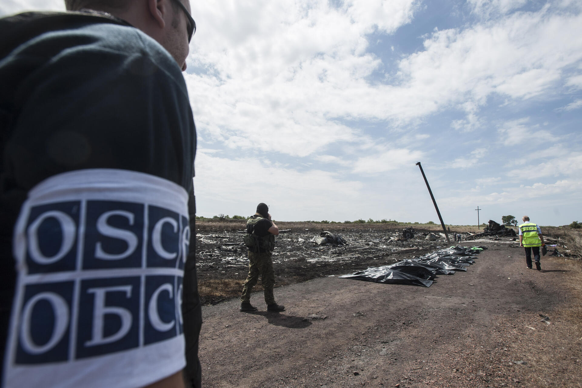 Inspectors in a field examine airplane debris, wearing uniforms with "OSCE" armbands.