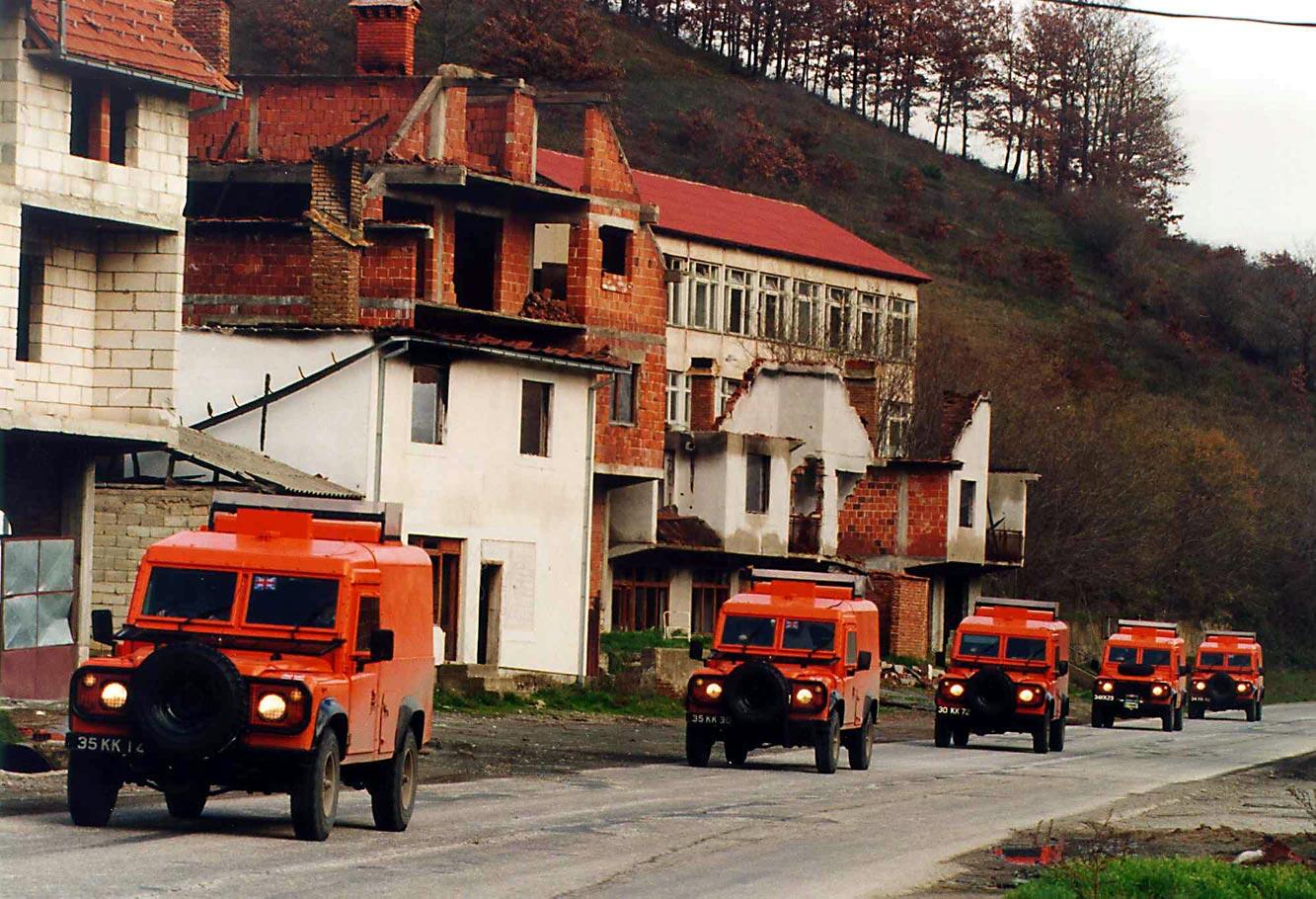 Convoy of orange armored vehicles driving along a war-torn street with damaged buildings.