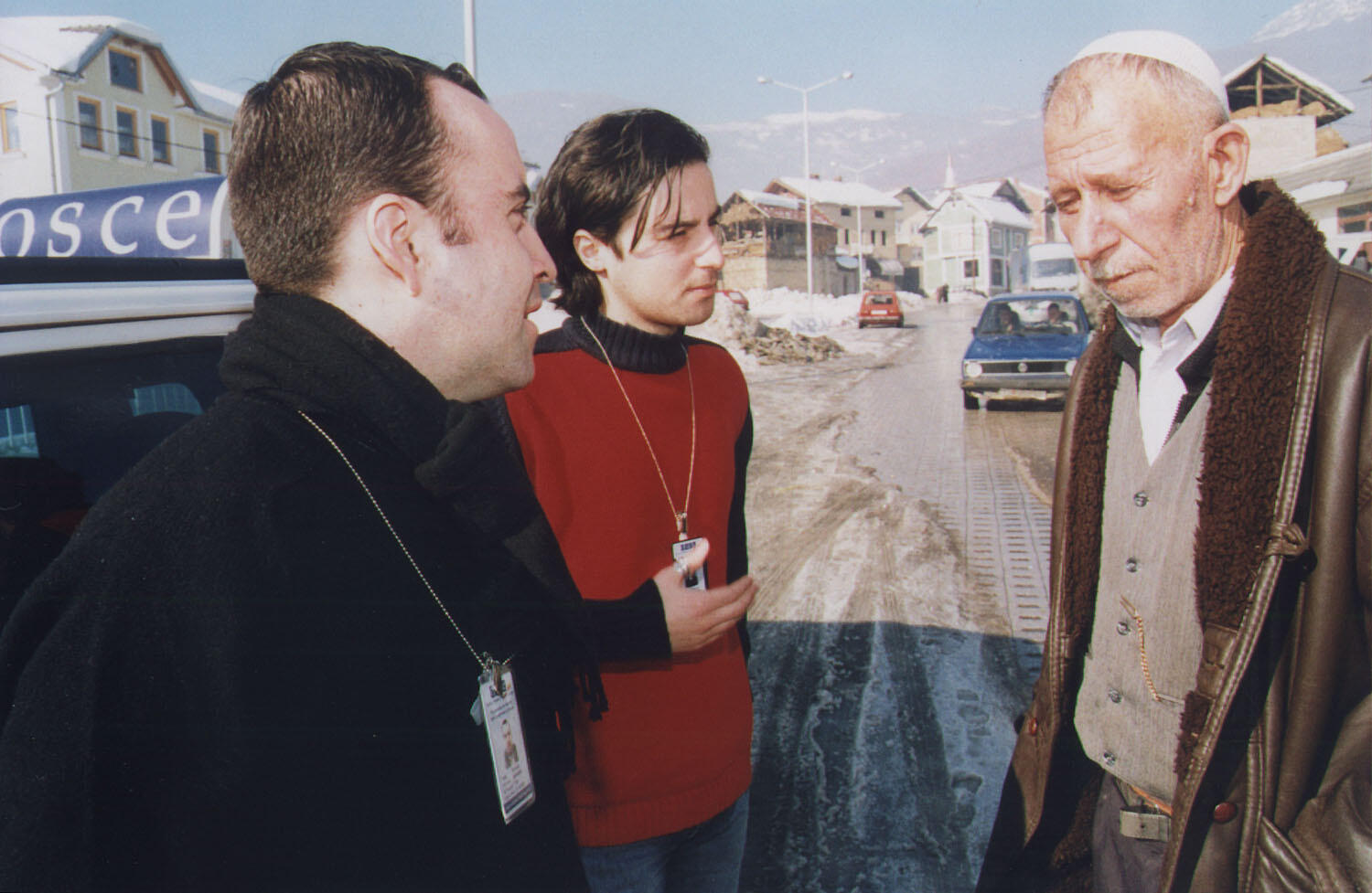 Two individuals with OSCE badges speaking to a man on a snowy street in a rural area.