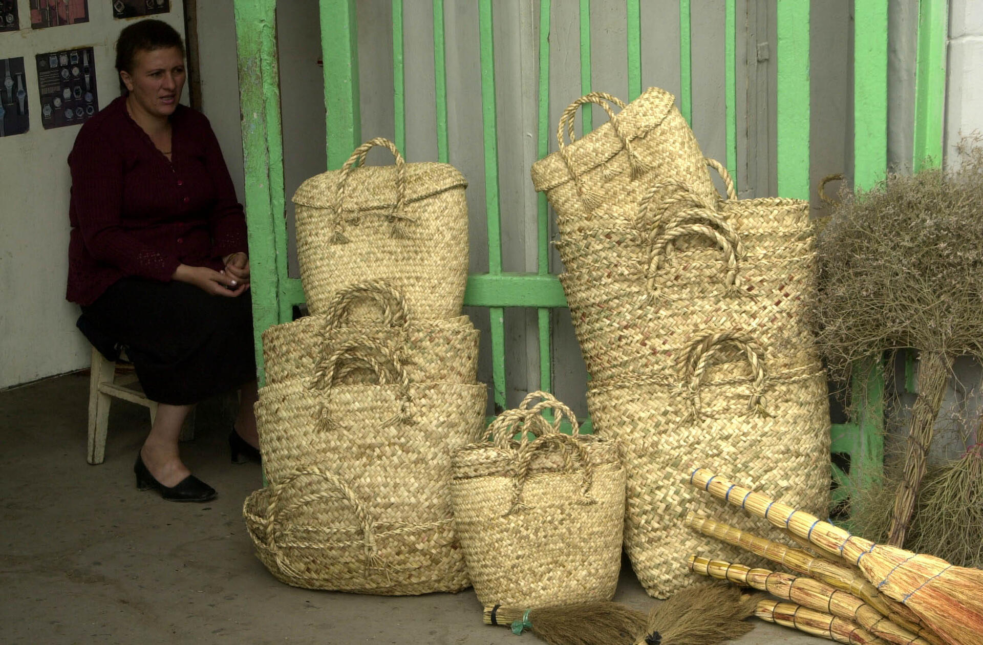 A woman sitting next to handmade woven baskets and brooms displayed for sale near a green gate.
