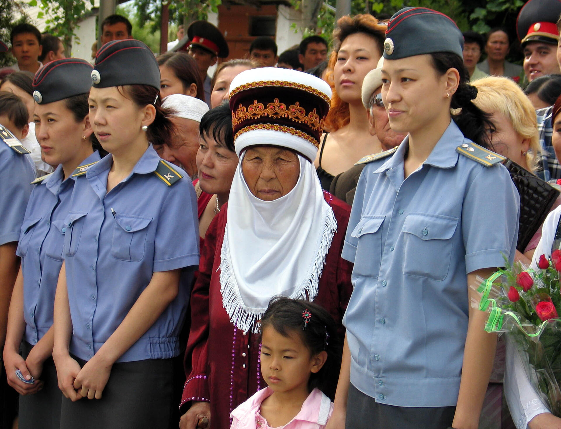 A group of women in uniform stands with a traditionally dressed older woman and a child at an outdoor event.