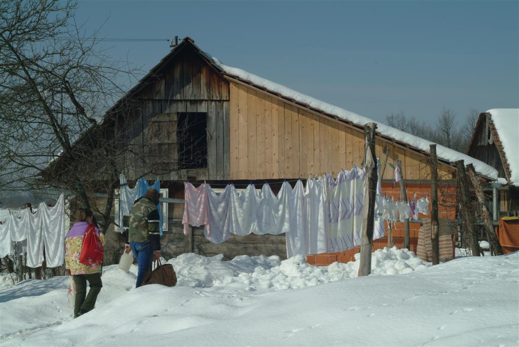 Two people stand by snowy clotheslines with laundry, in front of a rustic wooden barn.