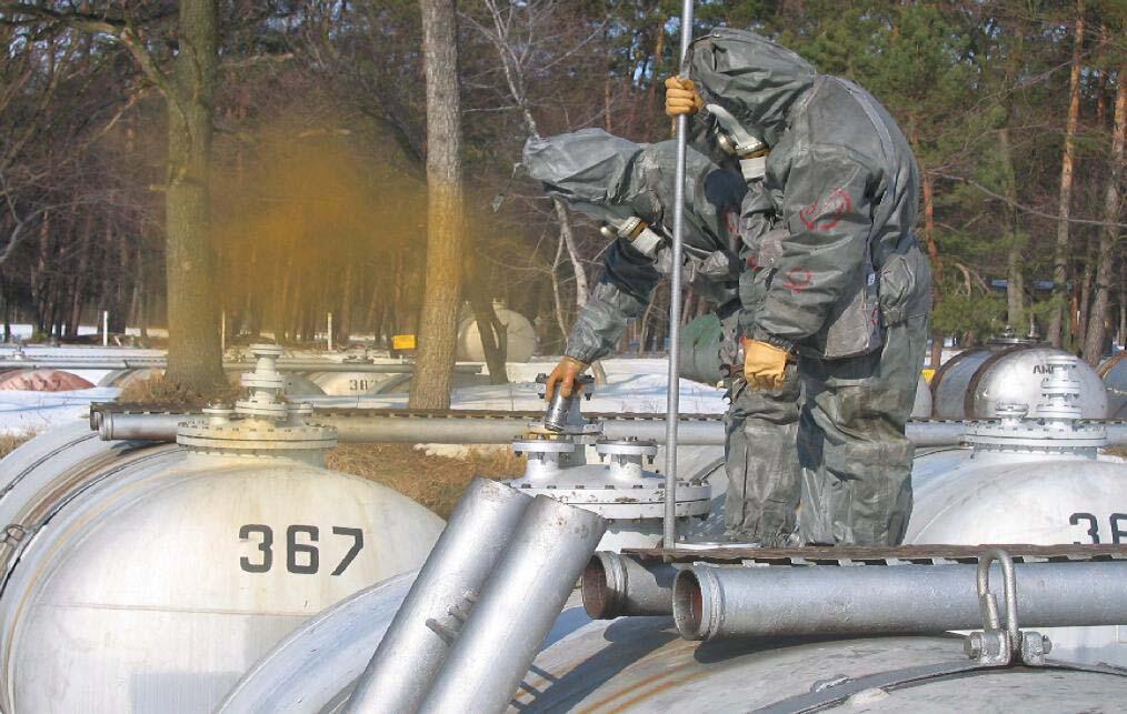 Workers in hazmat suits inspecting gas tanks with yellow fumes nearby.