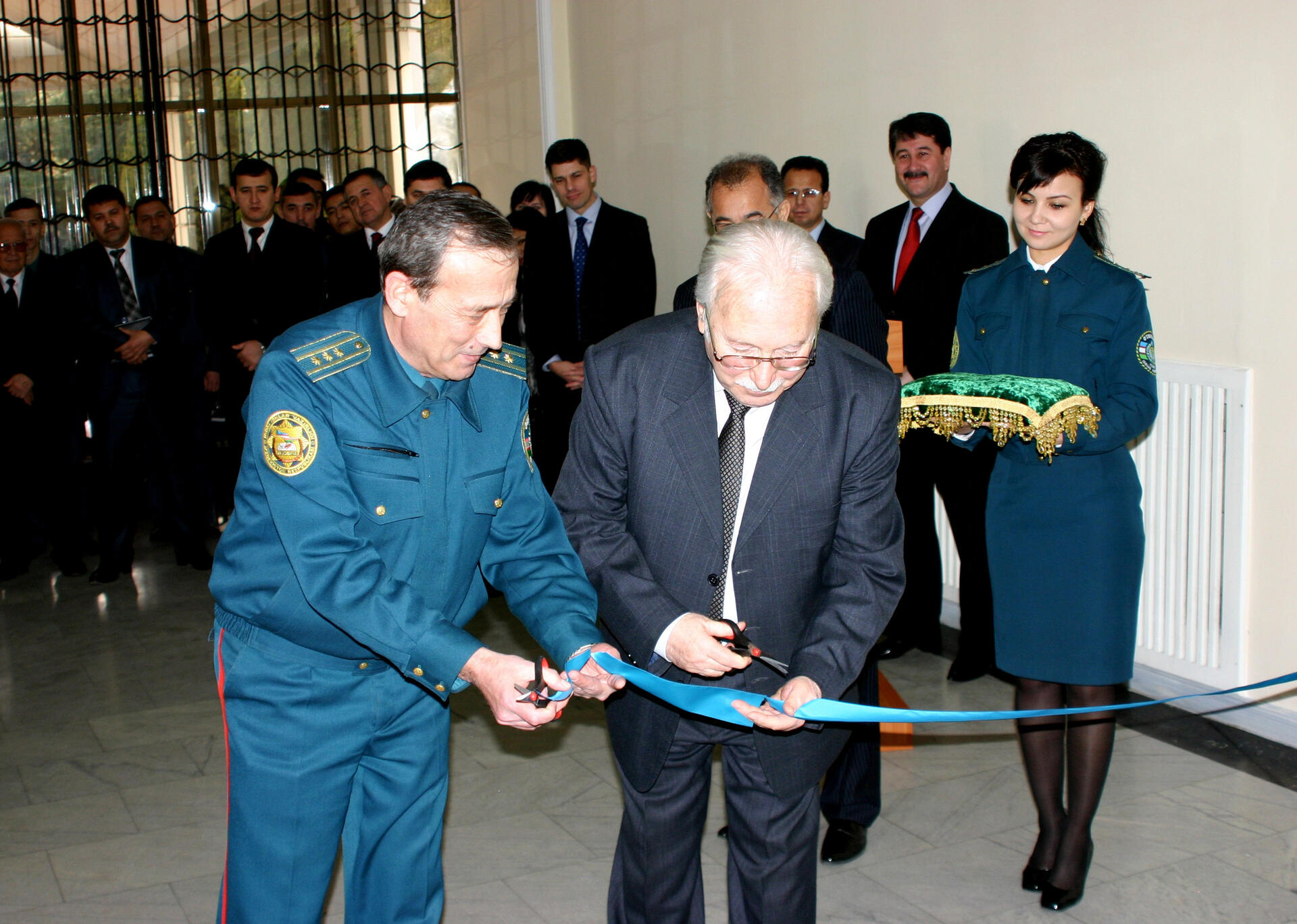 Two men cutting a ceremonial ribbon, surrounded by people, with a uniformed woman holding a decorative cloth.