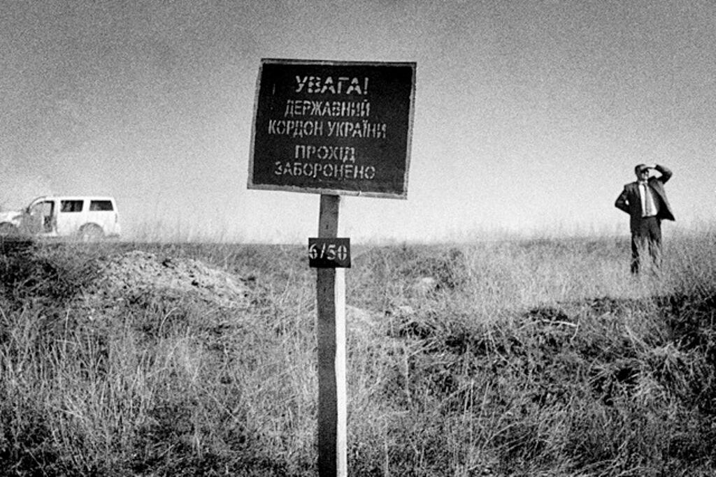 A man stands in a grassy field beside a sign, with a white SUV parked in the background.
