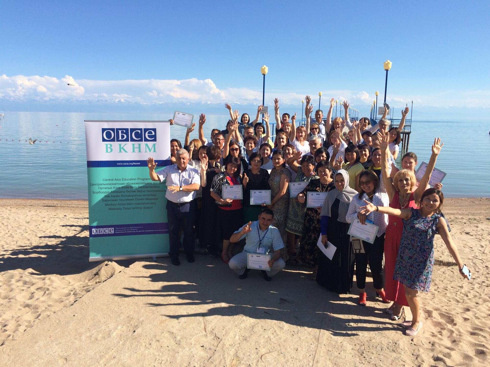 Group of people celebrating on a sunny beach, holding certificates with a scenic lake and mountains in the background.