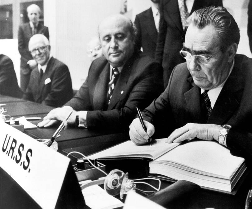 Black-and-white photograph of Leonid Brezhnev, General-Secretary of the Communist Party of the USSR, signing the Final Act at the Conference on Security and Co-operation in Europe, Helsinki, 1 August 1975. Brezhnev is focused on signing the document, with Turkey's Prime Minister Suleyman Demirel seated to his left, observing. A nameplate reading 'U.R.S.S.' is visible in the foreground, along with microphones and a folder.